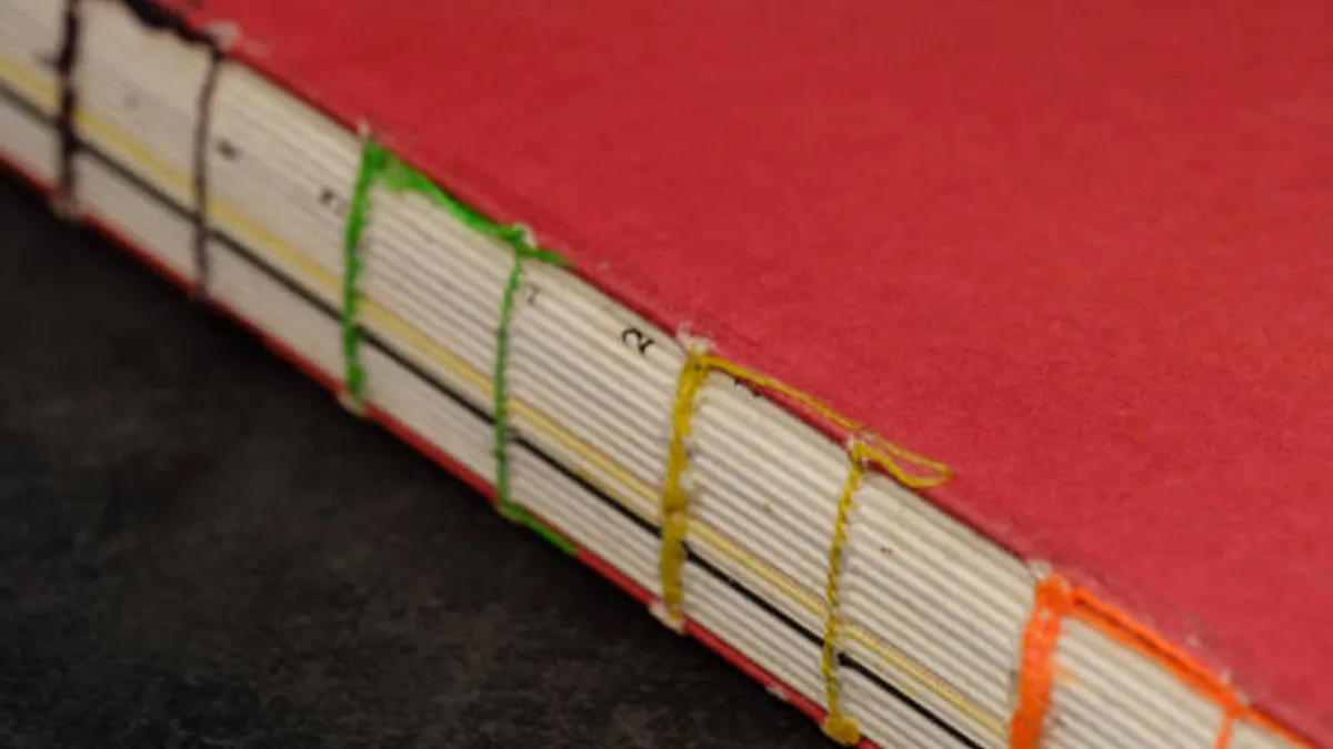 Edge of a red book sewn with black, green, yellow and orange thread through drilled holes in the covers