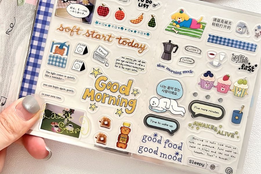 A hand holding a clear planner sticker page with kiss-cut morning themed stickers, including Good Morning lettering, a coffee pot, muffins and a sleeping figure